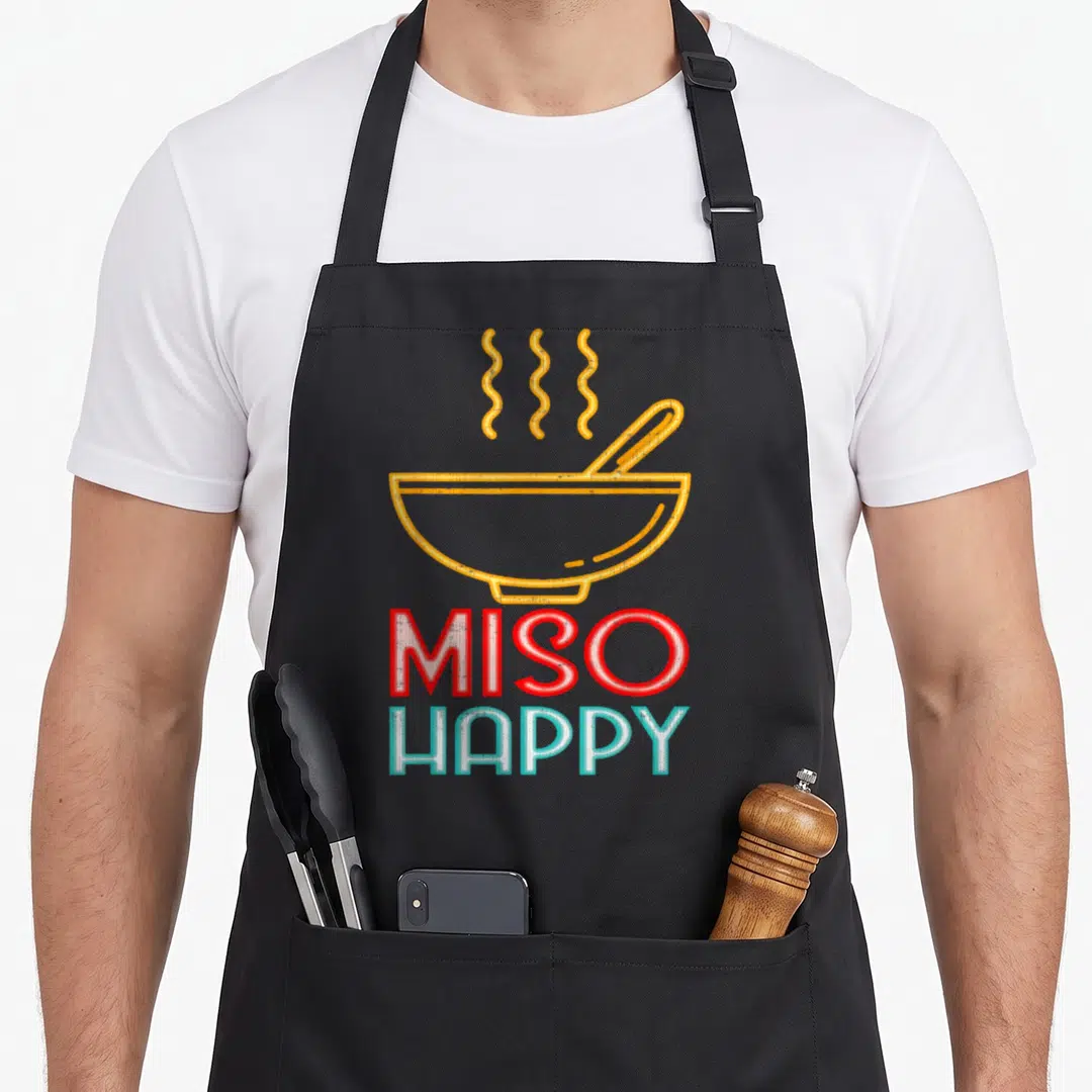 Miso food aprons - Cartoon soup bowl with steam above MISO HAPPY text in red and blue — shown on a black cotton canvas apron against a white background