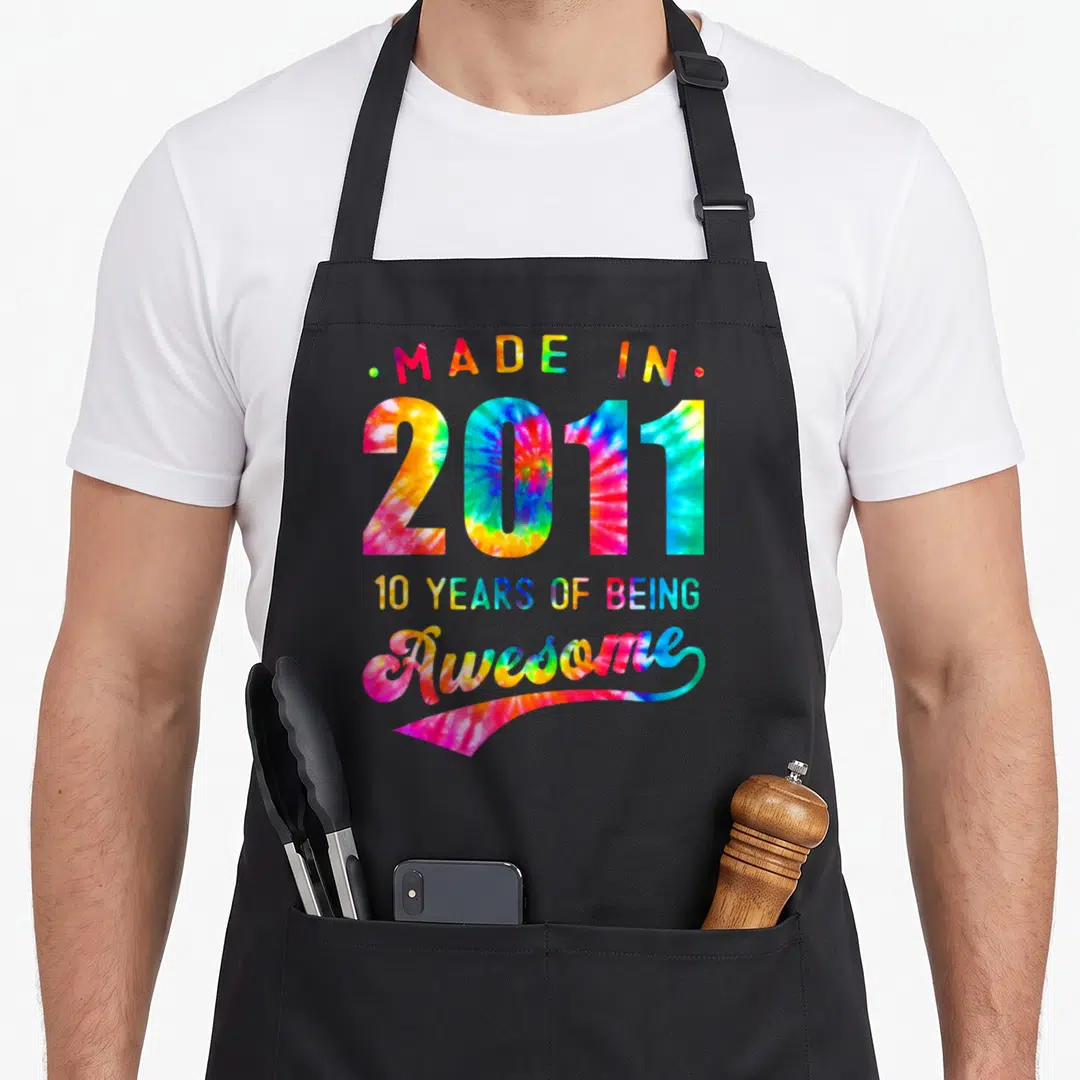 Birthday tie dye apron - Stacked tie-dye text reading Made in 2011 10 Years of Being Awesome in rainbow colors — shown on a black cotton canvas apron against a white background