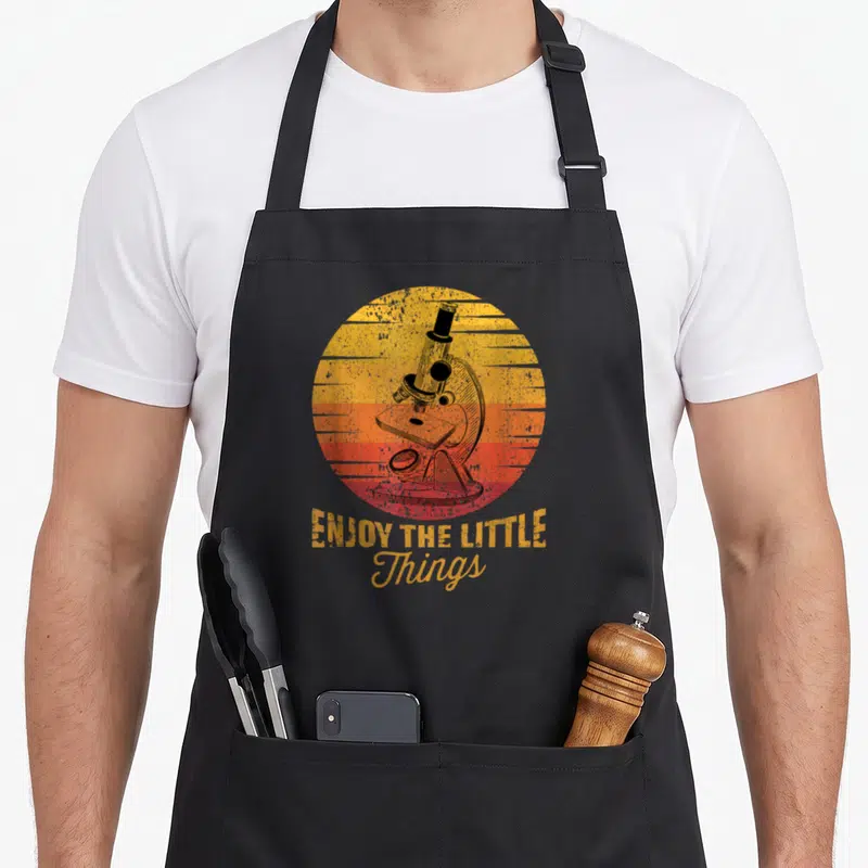 Biochemist science apron - Retro microscope inside striped sunset circle with stacked text ENJOY THE LITTLE Things — shown on a black cotton canvas apron against a white background