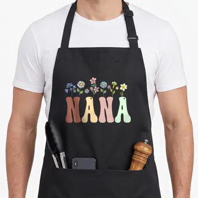 Nana wildflower floral apron - NANA text in brown yellow pink and green with wildflowers growing from letters — shown on a black cotton canvas apron against a white background
