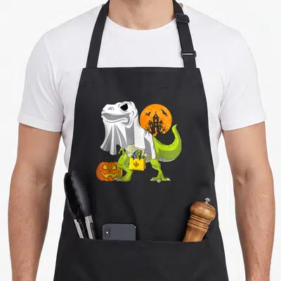 Ghost dinosaur halloween apron - Green dinosaur in white ghost sheet holding yellow bag next to orange pumpkin — shown on a black cotton canvas apron against a white background