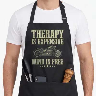 Funny motorcycle bike apron - Beige distressed text Therapy Is Expensive Wind Is Free above and below a motorcycle — shown on a black cotton canvas apron against a white background