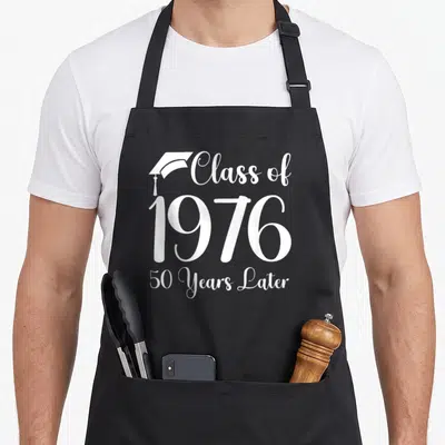Later class reunion apron - White Class of 1976 50 Years Later text with graduation cap icon — shown on a black cotton canvas apron against a white background