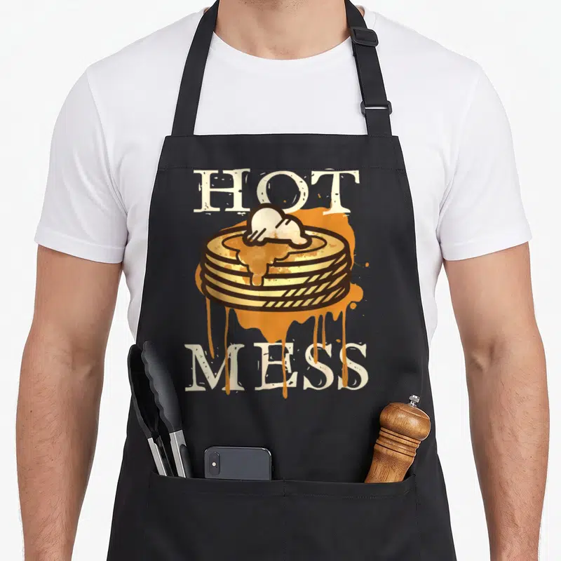 Hot pancake apron - Cartoon stack of pancakes with dripping syrup between HOT and MESS text — shown on a black cotton canvas apron against a white background