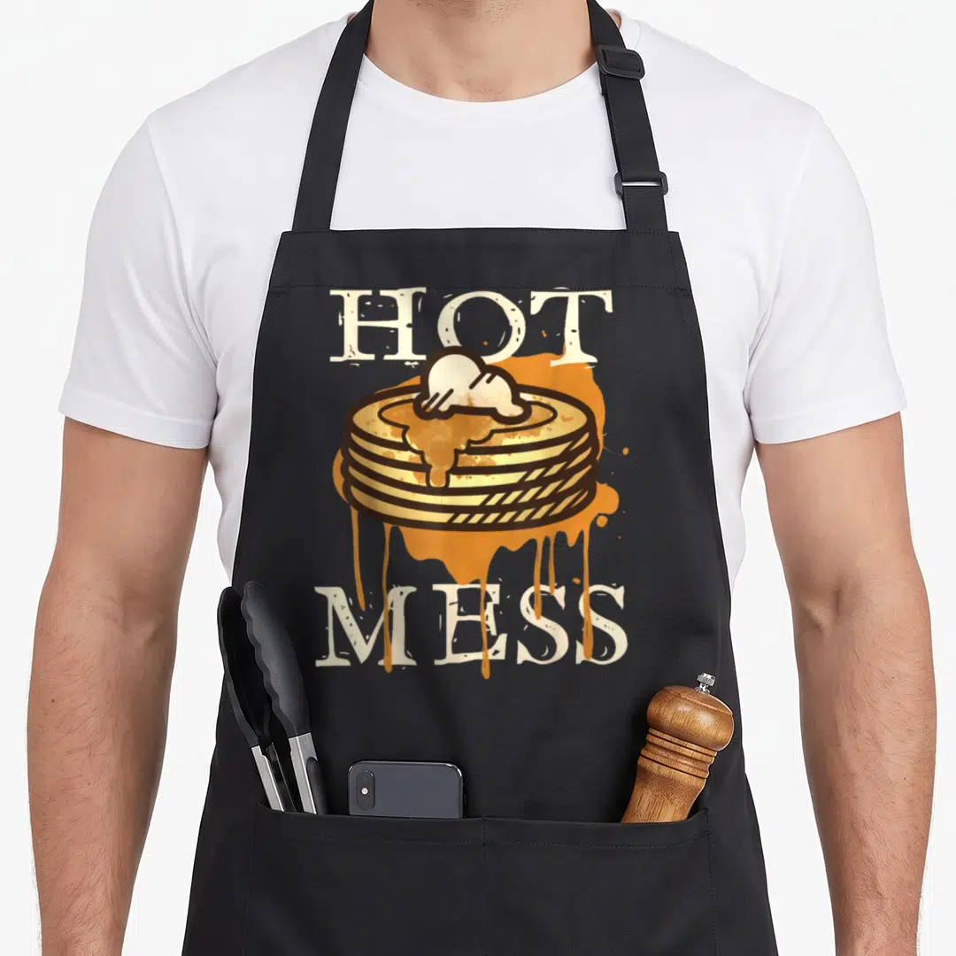 Hot pancake apron - Cartoon stack of pancakes with dripping syrup between HOT and MESS text — shown on a black cotton canvas apron against a white background
