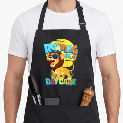 School daycare apron - Cartoon lion with sunglasses under arched text ROARING INTO DAYCARE above green grass — shown on a black cotton canvas apron against a white background