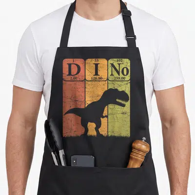 Dinosaur table apron - T-Rex silhouette over red orange and yellow periodic table cells spelling Dino — shown on a black cotton canvas apron against a white background