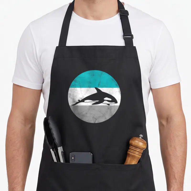 Killer whale orca apron - Grey orca silhouette on a distressed circle with teal, white, and grey horizontal bands — shown on a black cotton canvas apron against a white background
