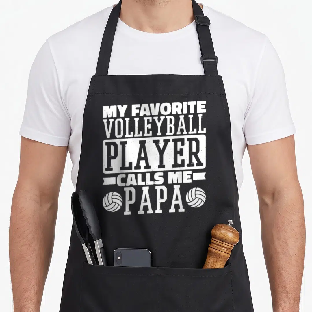 Volleyball papa apron - White distressed text reading My Favorite Volleyball Player Calls Me Papa flanked by two balls — shown on a black cotton canvas apron against a white background