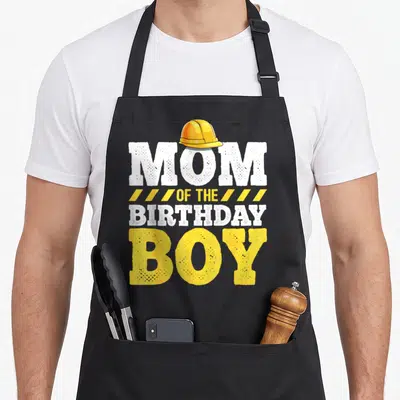 Hat construction apron - Yellow hard hat above distressed MOM OF THE BIRTHDAY BOY text with diagonal stripes — shown on a black cotton canvas apron against a white background