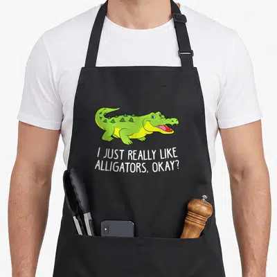 Crocodile alligator apron - Green cartoon alligator above white text reading I JUST REALLY LIKE ALLIGATORS OKAY — shown on a black cotton canvas apron against a white background