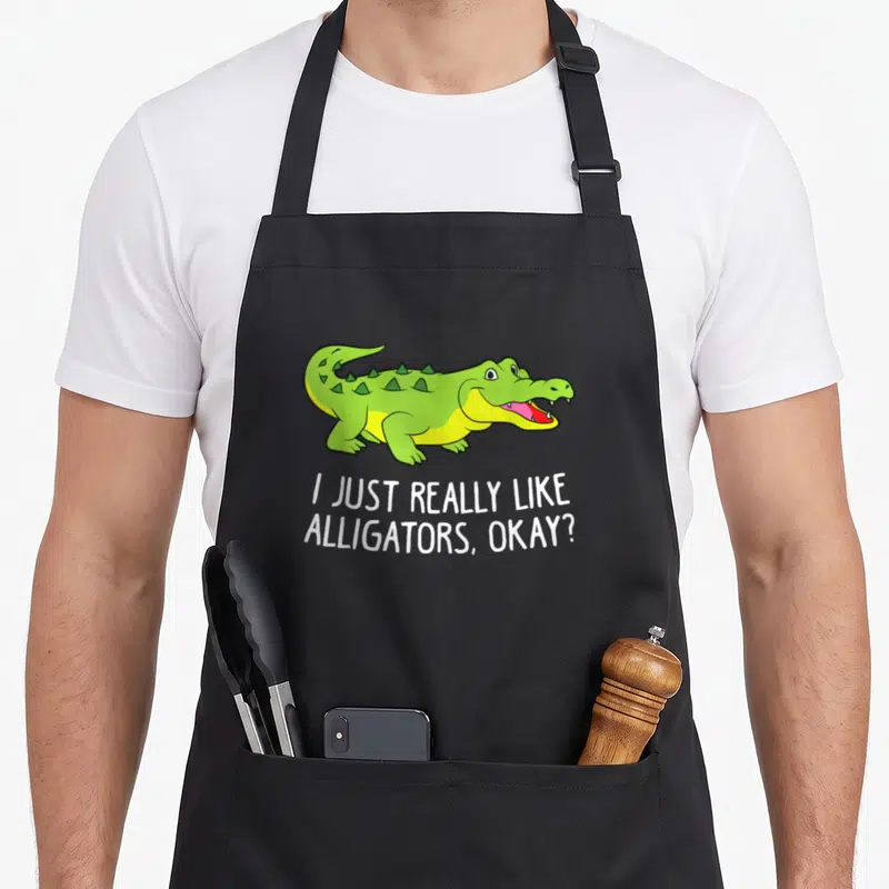 Crocodile alligator apron - Green cartoon alligator above white text reading I JUST REALLY LIKE ALLIGATORS OKAY — shown on a black cotton canvas apron against a white background