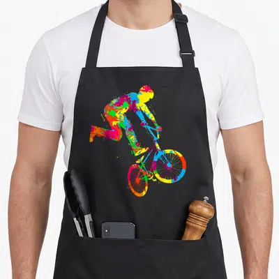 Bmx stunt bike apron - Multicolored splatter cartoon cyclist riding bicycle in yellow blue red green magenta — shown on a black cotton canvas apron against a white background
