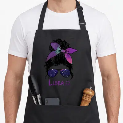 Zodiac constellation birthday apron - Woman's head with galaxy bow, constellation sunglasses, LIBRA Ω text in purple — shown on a black cotton canvas apron against a white background