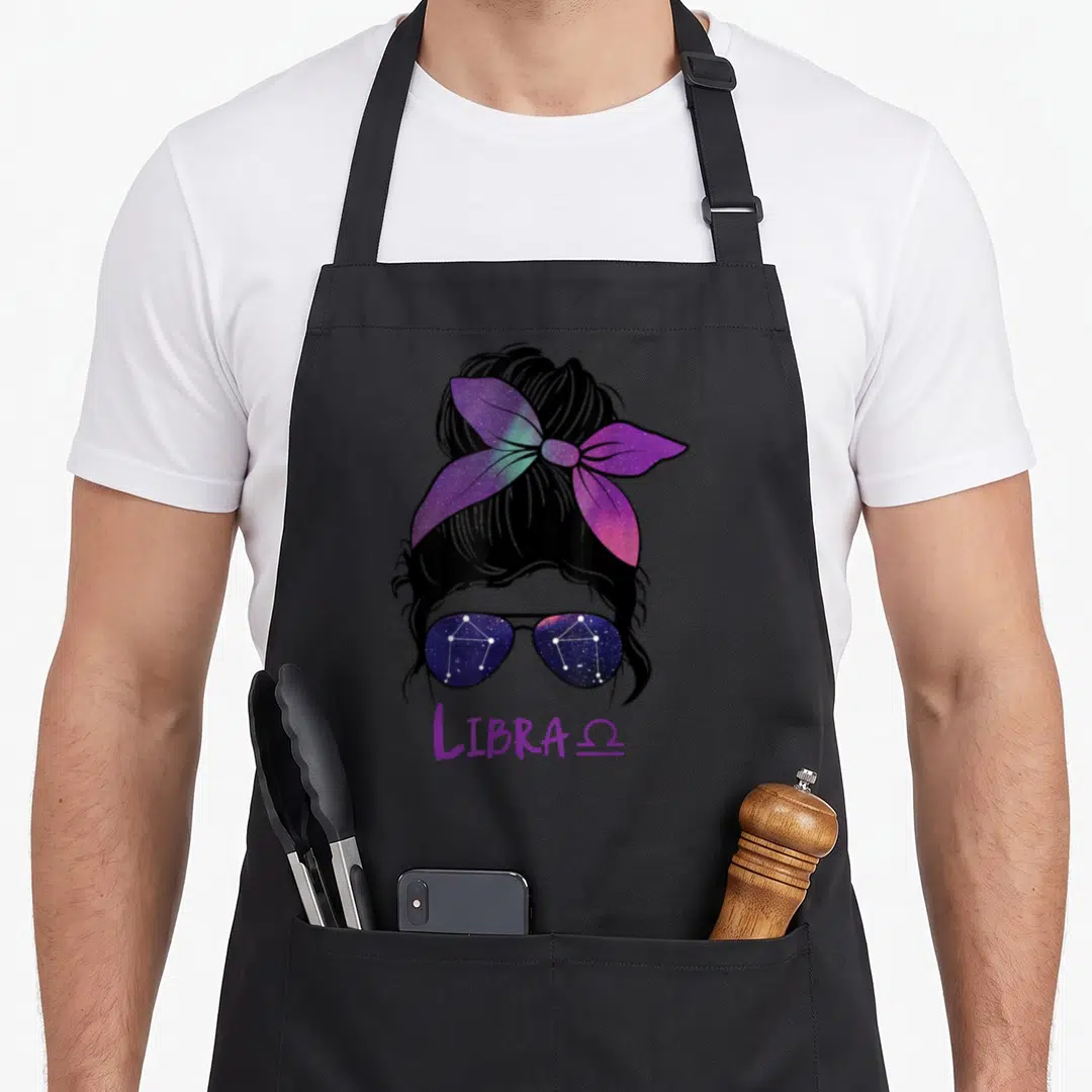 Zodiac constellation birthday apron - Woman's head with galaxy bow, constellation sunglasses, LIBRA Ω text in purple — shown on a black cotton canvas apron against a white background
