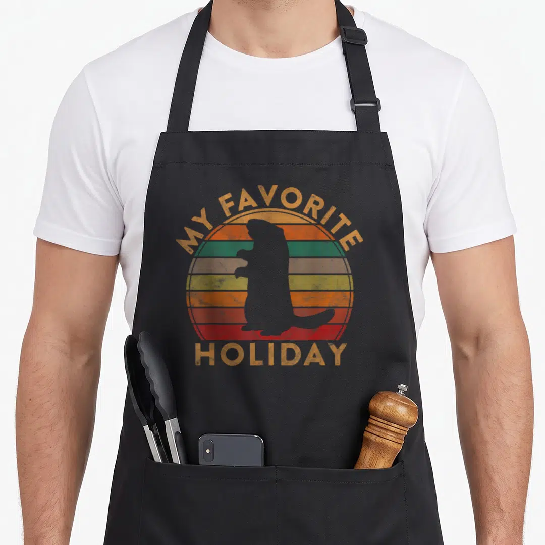 Ground holiday apron - Grey groundhog silhouette over orange green and red striped circle with arched text — shown on a black cotton canvas apron against a white background
