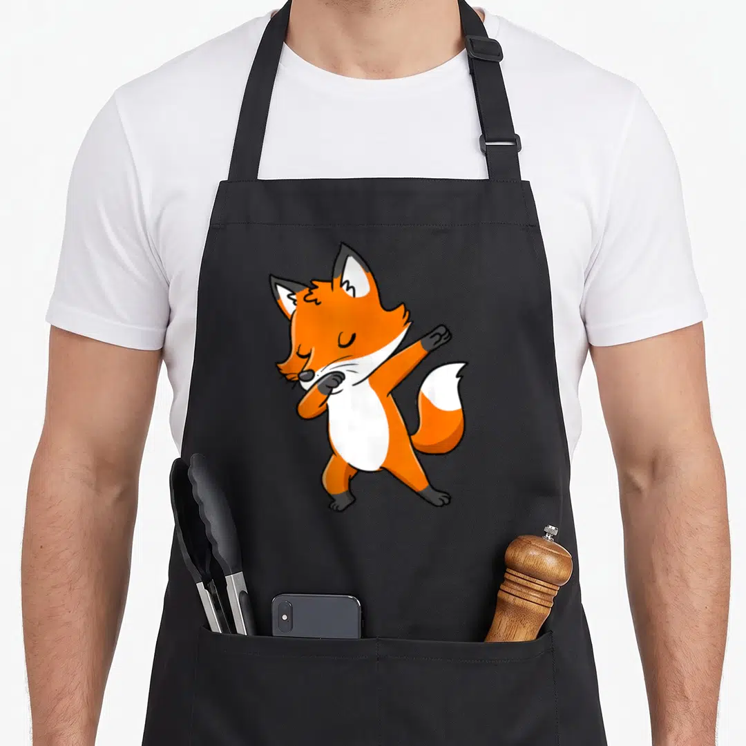Dabbing fox print apron - Cartoon orange and white fox doing a dab pose with closed eyes — shown on a black cotton canvas apron against a white background