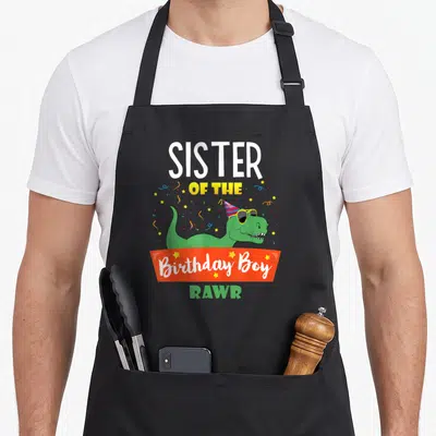 Sister birthday dinosaur apron - Green cartoon dinosaur in sunglasses and party hat with SISTER OF THE Birthday Boy — shown on a black cotton canvas apron against a white background