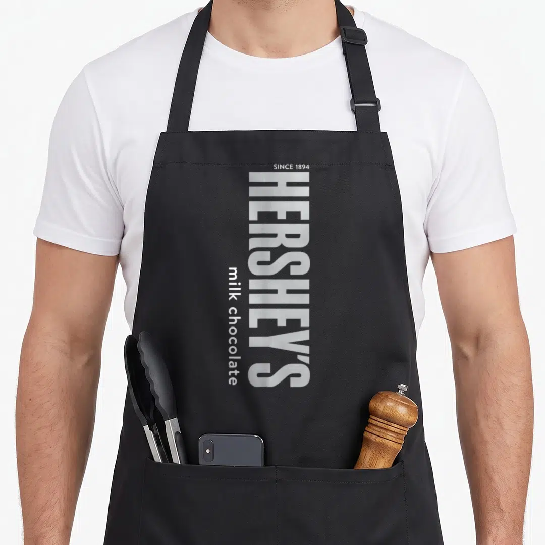 Hershey milk aprons - Vertical white HERSHEY'S text with SINCE 1894 and milk chocolate labels — shown on a black cotton canvas apron against a white background