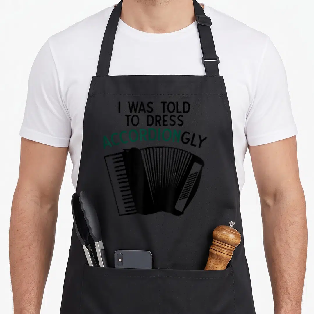 Accordion joke apron - Text I WAS TOLD TO DRESS ACCORDIONGLY above black and white cartoon accordion illustration — shown on a black cotton canvas apron against a white background