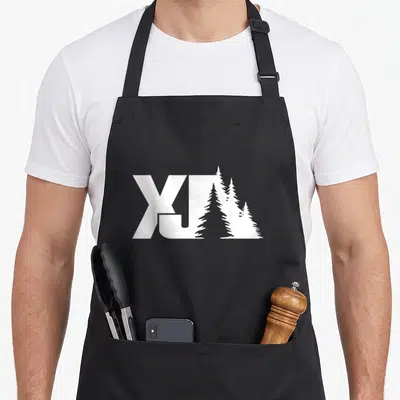Rock road apron - White X and J letters with three pine tree silhouettes replacing the J stem — shown on a black cotton canvas apron against a white background