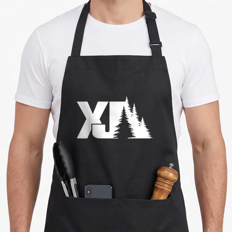 Rock road apron - White X and J letters with three pine tree silhouettes replacing the J stem — shown on a black cotton canvas apron against a white background