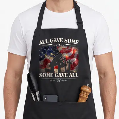 Gave veteran memorial apron - Soldier silhouette kneeling by cross and rifle over flag and fireworks with All Gave Some — shown on a black cotton canvas apron against a white background