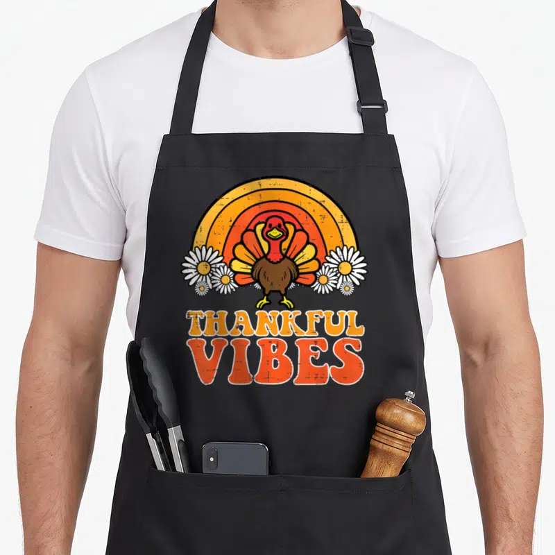 Turkey thankful apron - Cartoon turkey in front of rainbow and daisies with stacked THANKFUL VIBES text in orange — shown on a black cotton canvas apron against a white background
