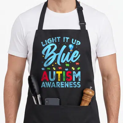 Blue light up apron - Light It Up Blue text, bulb with puzzle piece, colorful scattered pieces, Autism Awareness — shown on a black cotton canvas apron against a white background