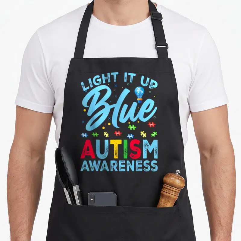 Blue light up apron - Light It Up Blue text, bulb with puzzle piece, colorful scattered pieces, Autism Awareness — shown on a black cotton canvas apron against a white background