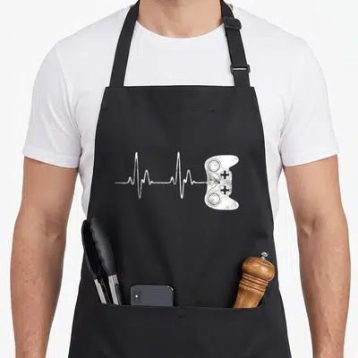 Heartbeat gaming apron - White distressed game controller connected to a heartbeat EKG line on black fabric — shown on a black cotton canvas apron against a white background