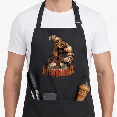 Juggernaut x apron - Realistic character crashing through ground with arched JUGGERNAUT text in brown and orange — shown on a black cotton canvas apron against a white background