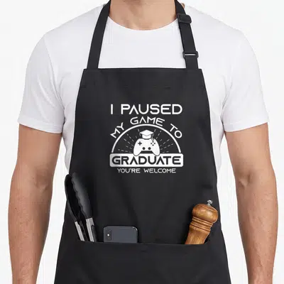 Graduate game aprons - White stacked text I paused my game to graduate above a controller wearing a cap — shown on a black cotton canvas apron against a white background