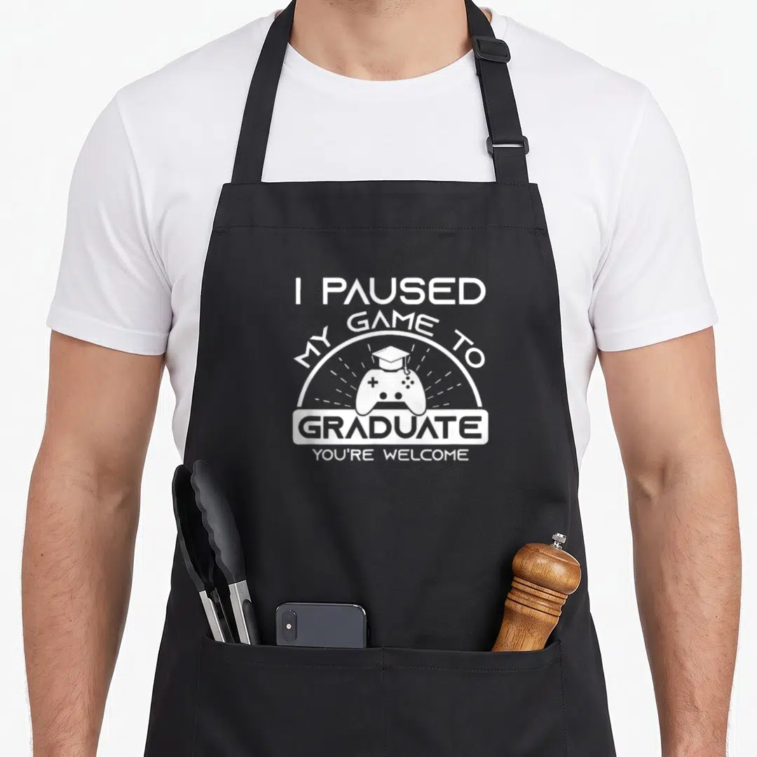 Graduate game aprons - White stacked text I paused my game to graduate above a controller wearing a cap — shown on a black cotton canvas apron against a white background