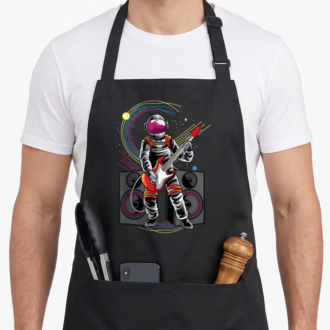 Astronaut playing guitar apron - Cartoon astronaut holding red electric guitar in front of two speakers with yellow — shown on a black cotton canvas apron against a white background