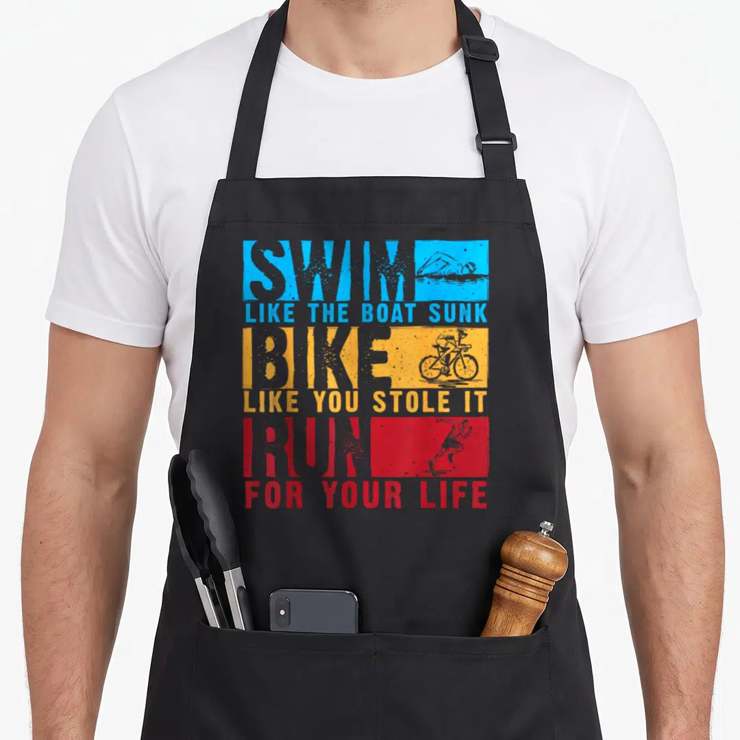 Boat bike apron - Swim bike run triathlon quote in blue orange red distressed blocks with athlete silhouettes — shown on a black cotton canvas apron against a white background