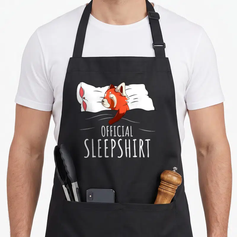 Sleepshirt panda apron - Red panda sleeping on white pillow under grey blanket with official sleepshirt text — shown on a black cotton canvas apron against a white background