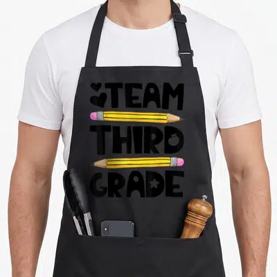 Grade 3rd teacher apron - Stacked TEAM THIRD GRADE text separated by two yellow pencils with pink erasers and black — shown on a black cotton canvas apron against a white background