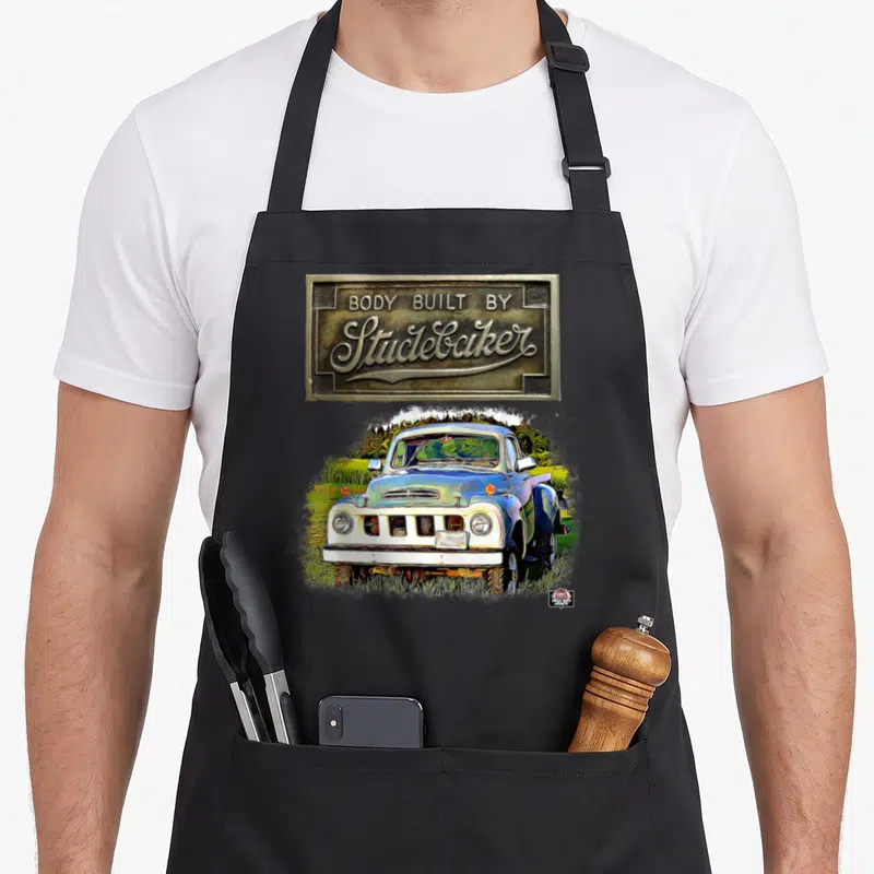 Studebaker truck apron - Brass emblem reading Body Built By Studebaker above blue and white vintage pickup in grass — shown on a black cotton canvas apron against a white background