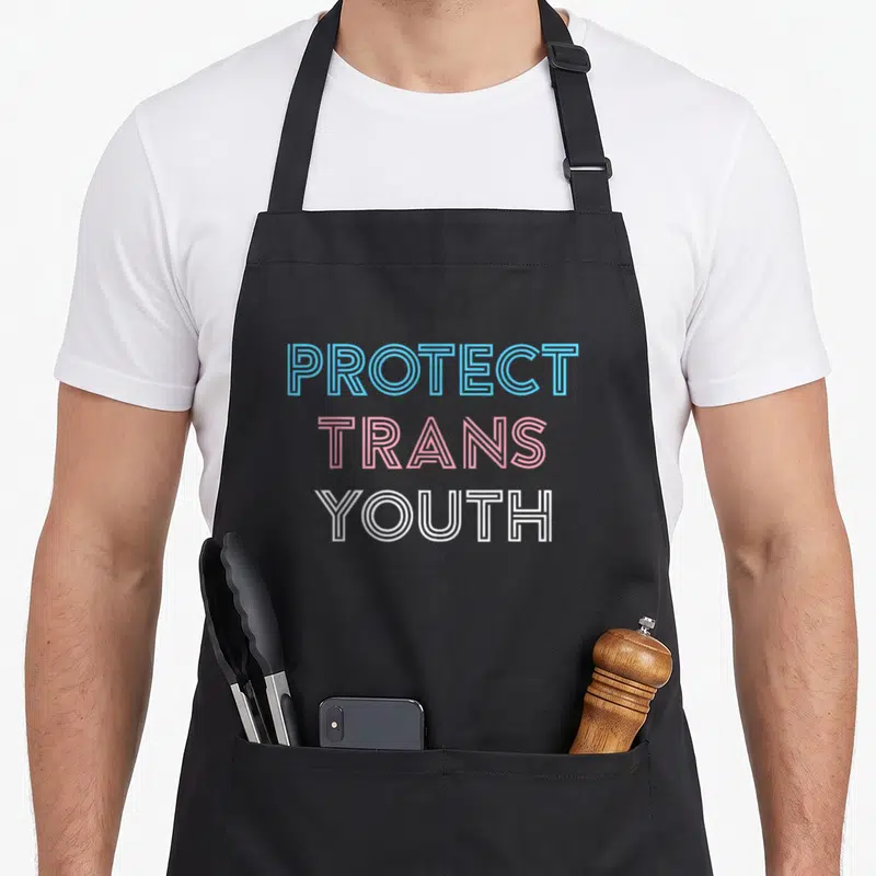 Protect pride apron - PROTECT TRANS YOUTH text in blue pink and white stacked typography — shown on a black cotton canvas apron against a white background