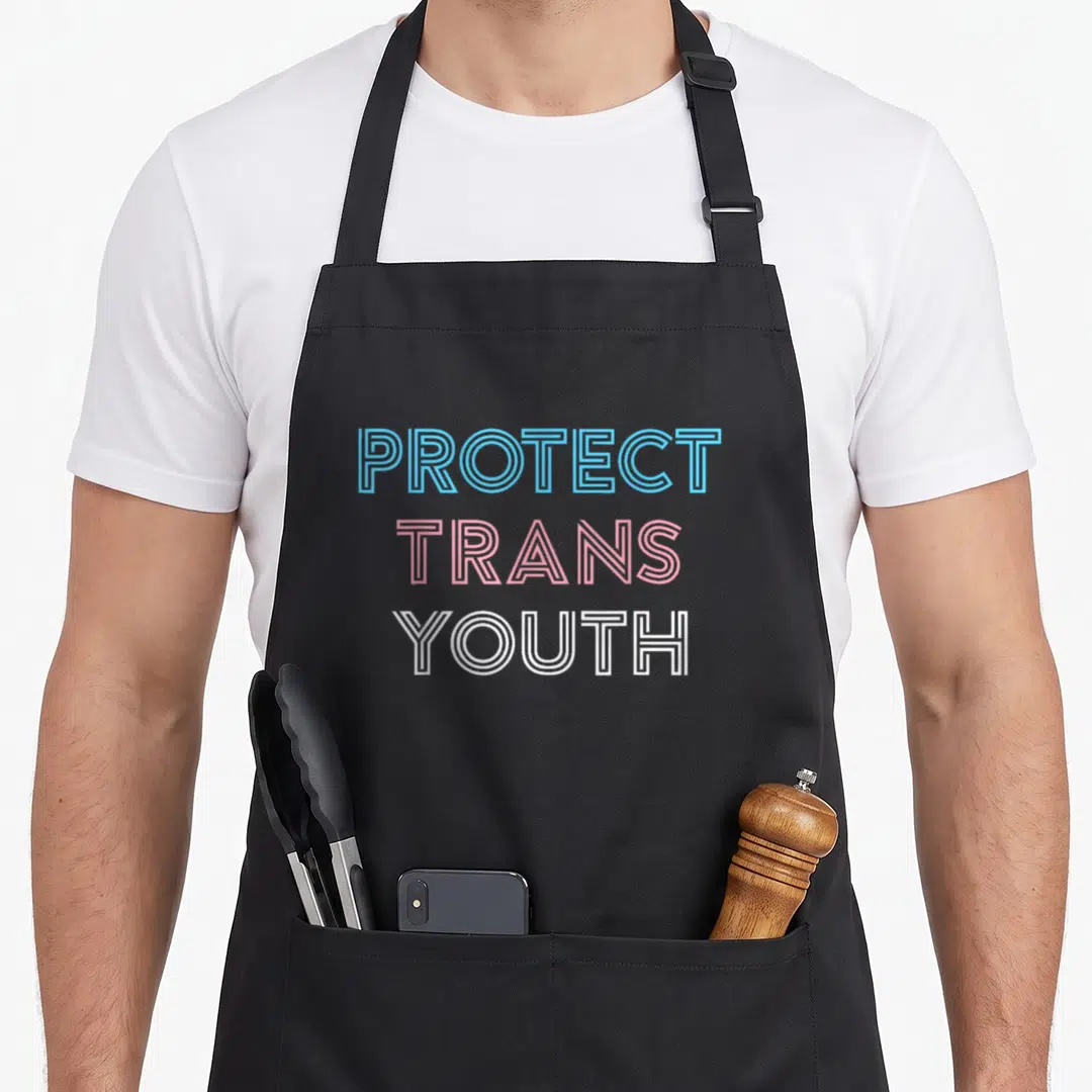 Protect pride apron - PROTECT TRANS YOUTH text in blue pink and white stacked typography — shown on a black cotton canvas apron against a white background