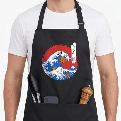 Wave cookies apron - Cartoon blue wave with eyes eating brown cookies in front of red sun with kanji banner — shown on a black cotton canvas apron against a white background