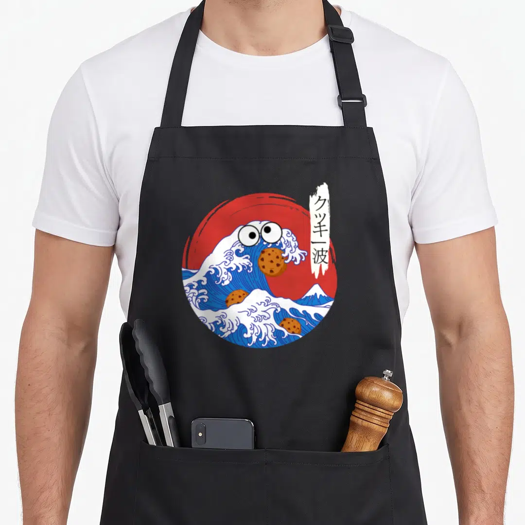 Wave cookies apron - Cartoon blue wave with eyes eating brown cookies in front of red sun with kanji banner — shown on a black cotton canvas apron against a white background