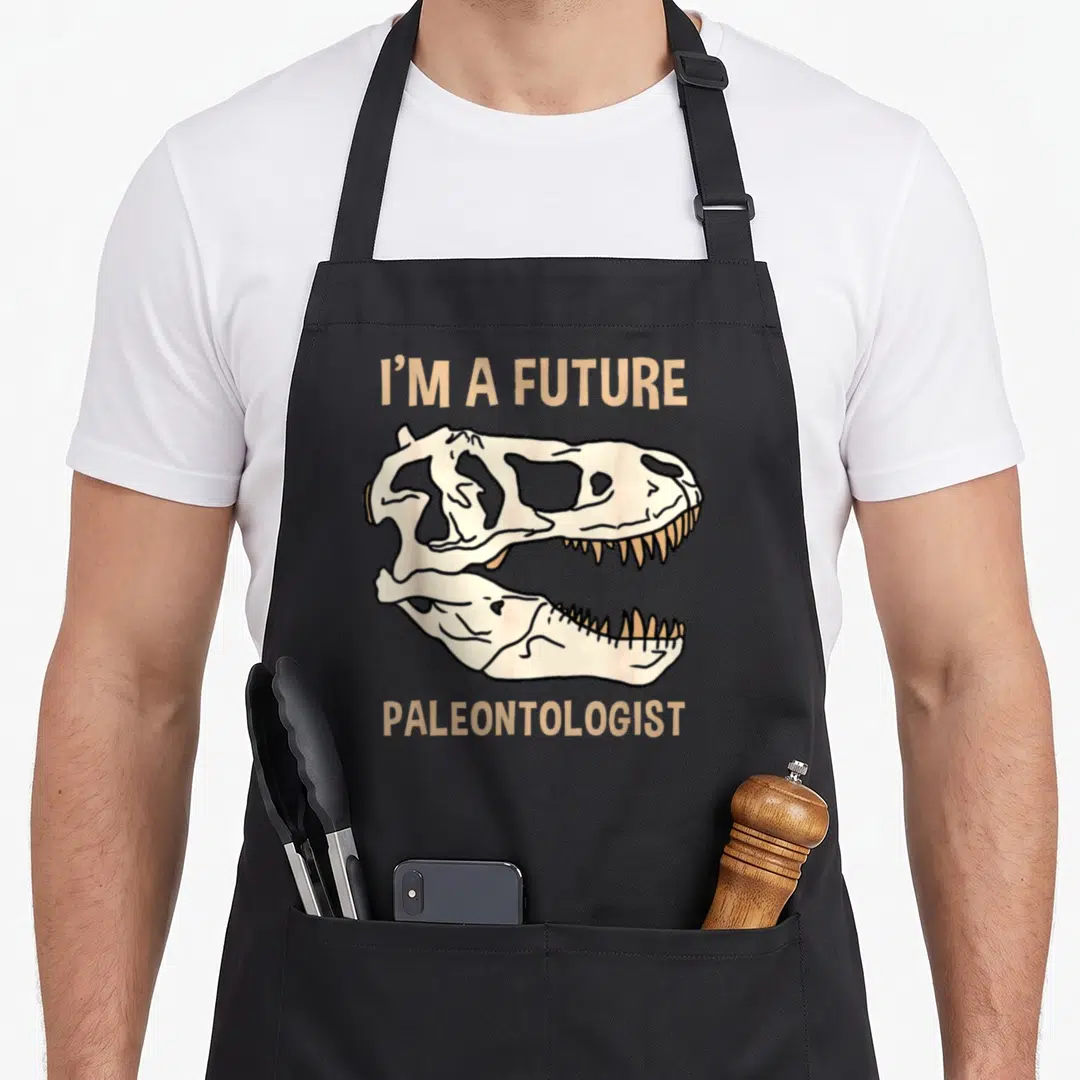 Future paleontology hobby apron - Cartoon dinosaur skull with tan text I'M A FUTURE PALEONTOLOGIST above and below — shown on a black cotton canvas apron against a white background