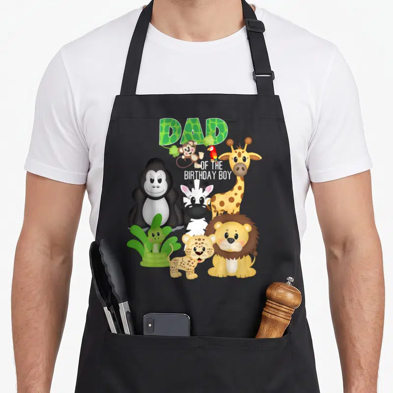 Jungle safari zoo birthday apron - DAD OF THE BIRTHDAY BOY text over cartoon gorilla lion zebra giraffe monkey leopard — shown on a black cotton canvas apron against a white background