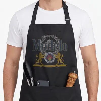 Modelo lager apron - Grey gold and white Modelo logo with two lions wheat stalks and 1925 banner — shown on a black cotton canvas apron against a white background