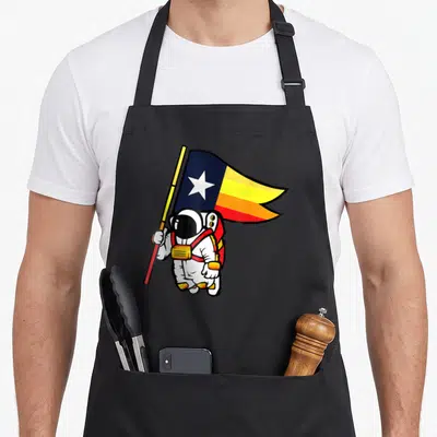 Astronaut space houston apron - Cartoon astronaut holding tricolor flag with white star in blue yellow and red — shown on a black cotton canvas apron against a white background
