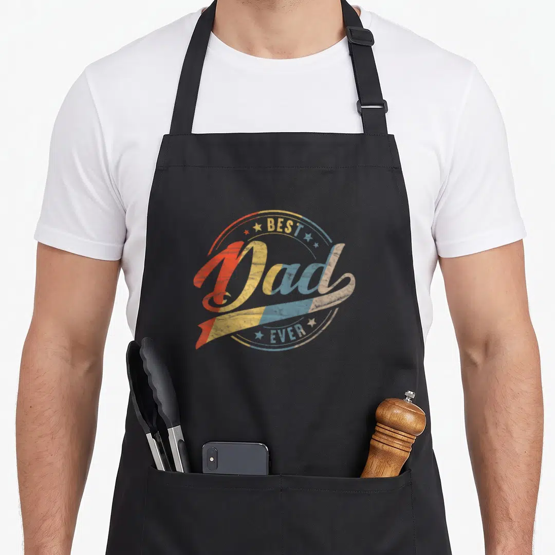 Cute father daddy aprons - Retro BEST Dad EVER text in red yellow blue with stars in circular composition — shown on a black cotton canvas apron against a white background