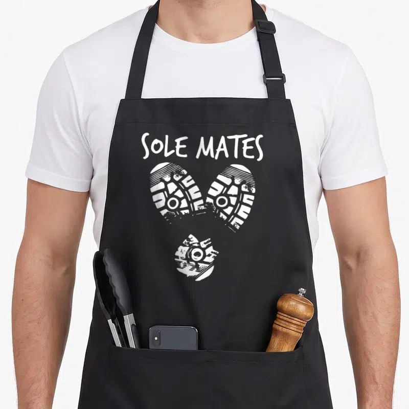 Mate sole apron - SOLE MATES text above three white shoe prints arranged in a heart shape — shown on a black cotton canvas apron against a white background