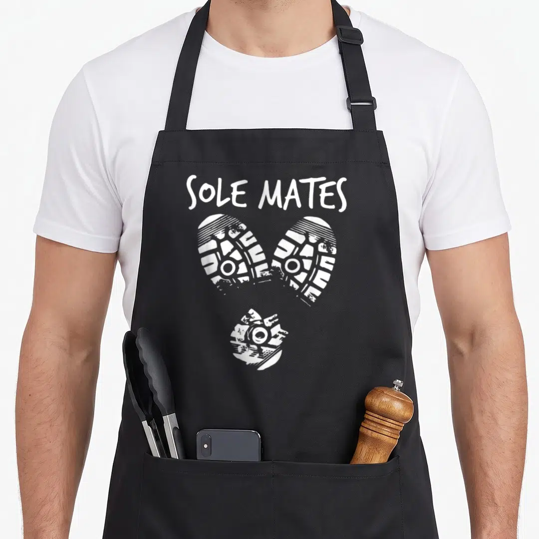 Mate sole apron - SOLE MATES text above three white shoe prints arranged in a heart shape — shown on a black cotton canvas apron against a white background